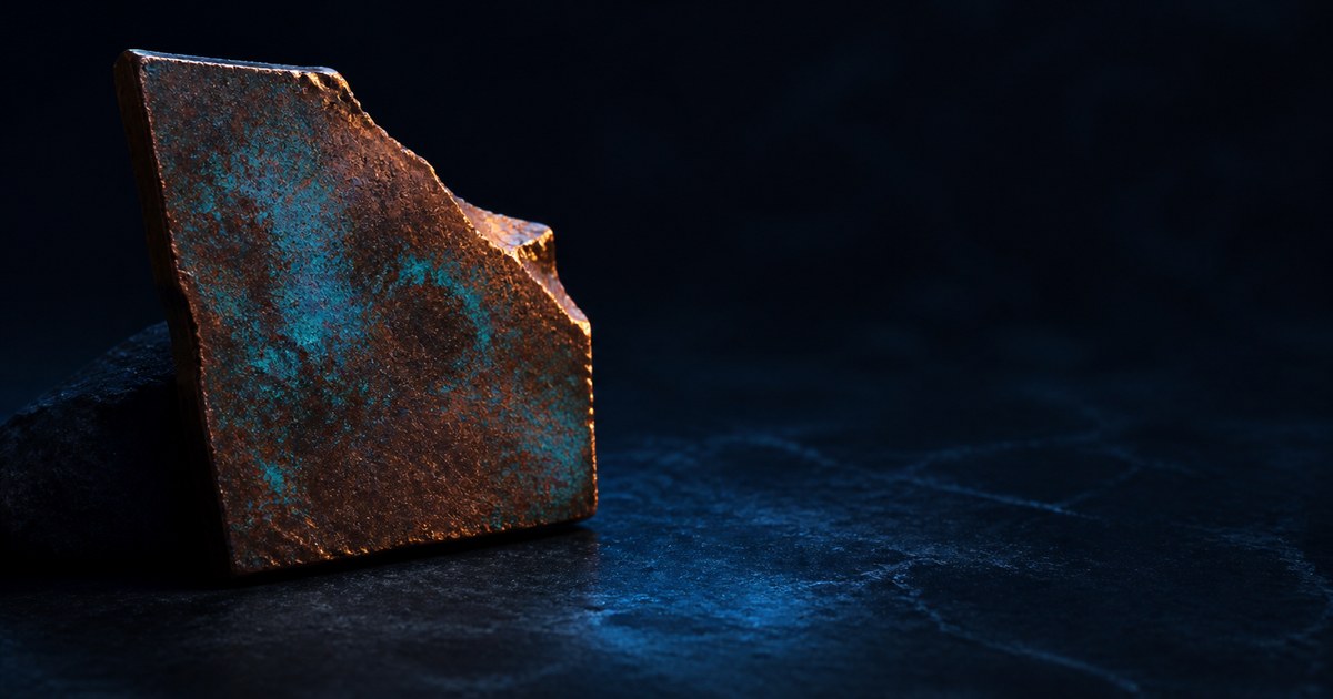 A blue-green oxidized copper fragment standing on dark stone with warm metal exposed along one chipped edge